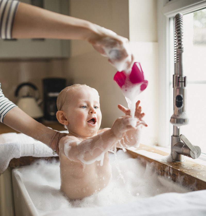 A mother bathes her baby boy in the kitchen sink, he is playing with the bubbles the mother is pouring on to him.